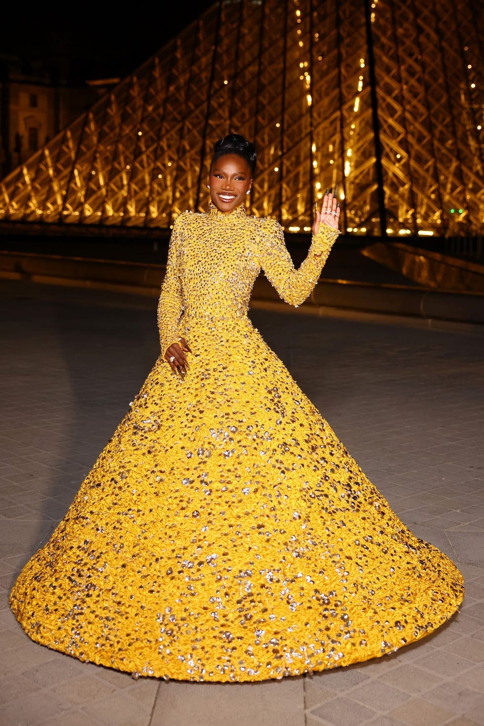 a person in a dazzling yellow dress with intricate detailing posing against a backdrop of illuminated architecture