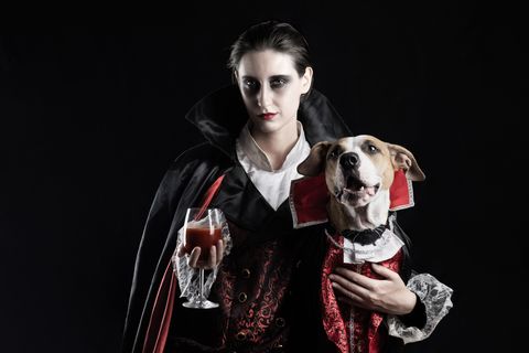 young female with glass of red drink and her pet puppy dressed up in same dracula costume