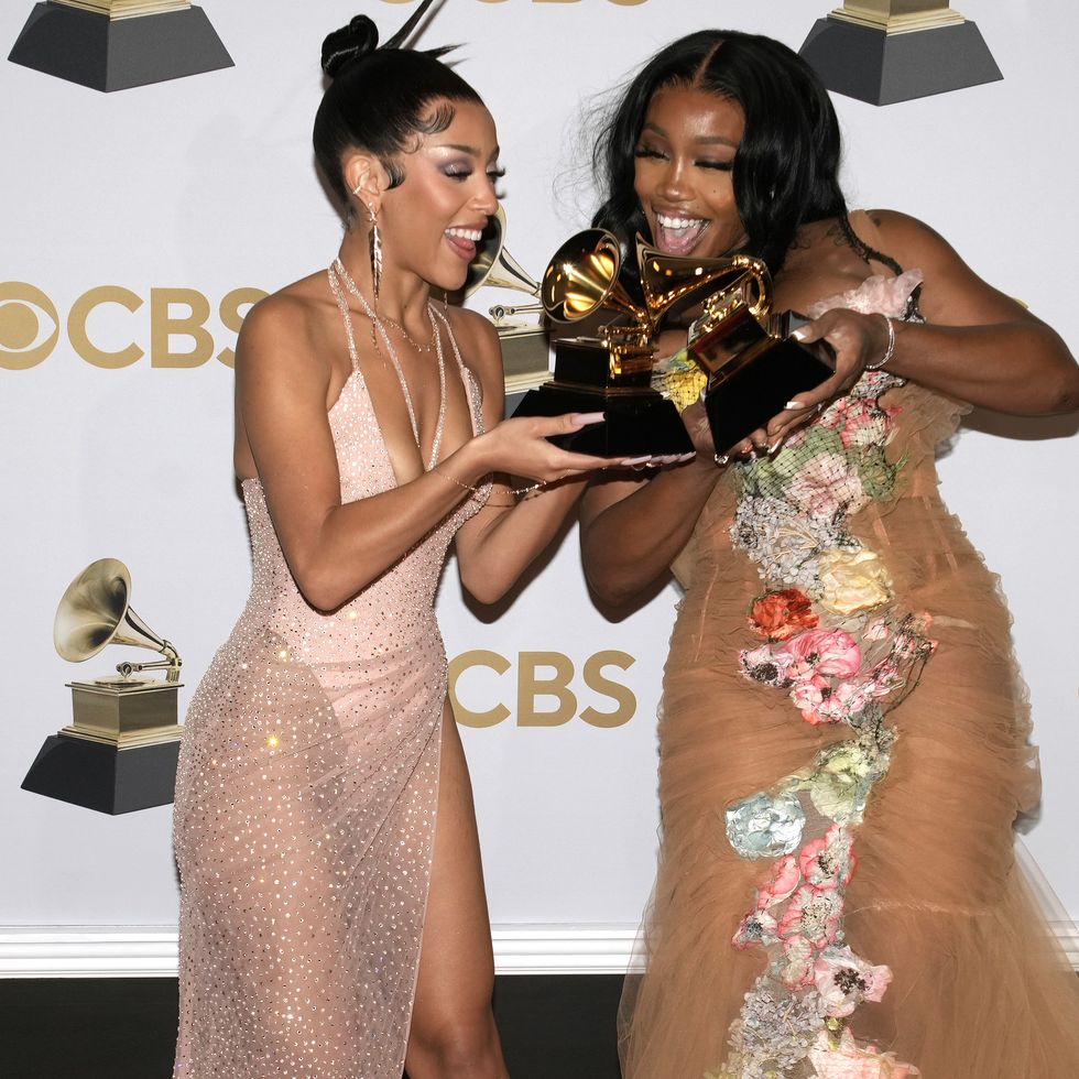 doja cat and sza each hold a grammy trophy and smile while standing together, both women wear dresses