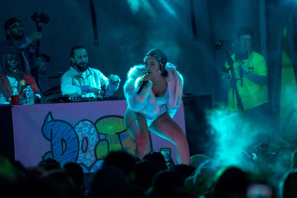 doja cat sings into a microphone she holds in one hand as she leans forward and stands on a stage, a dj sits at a table with equipment behind her