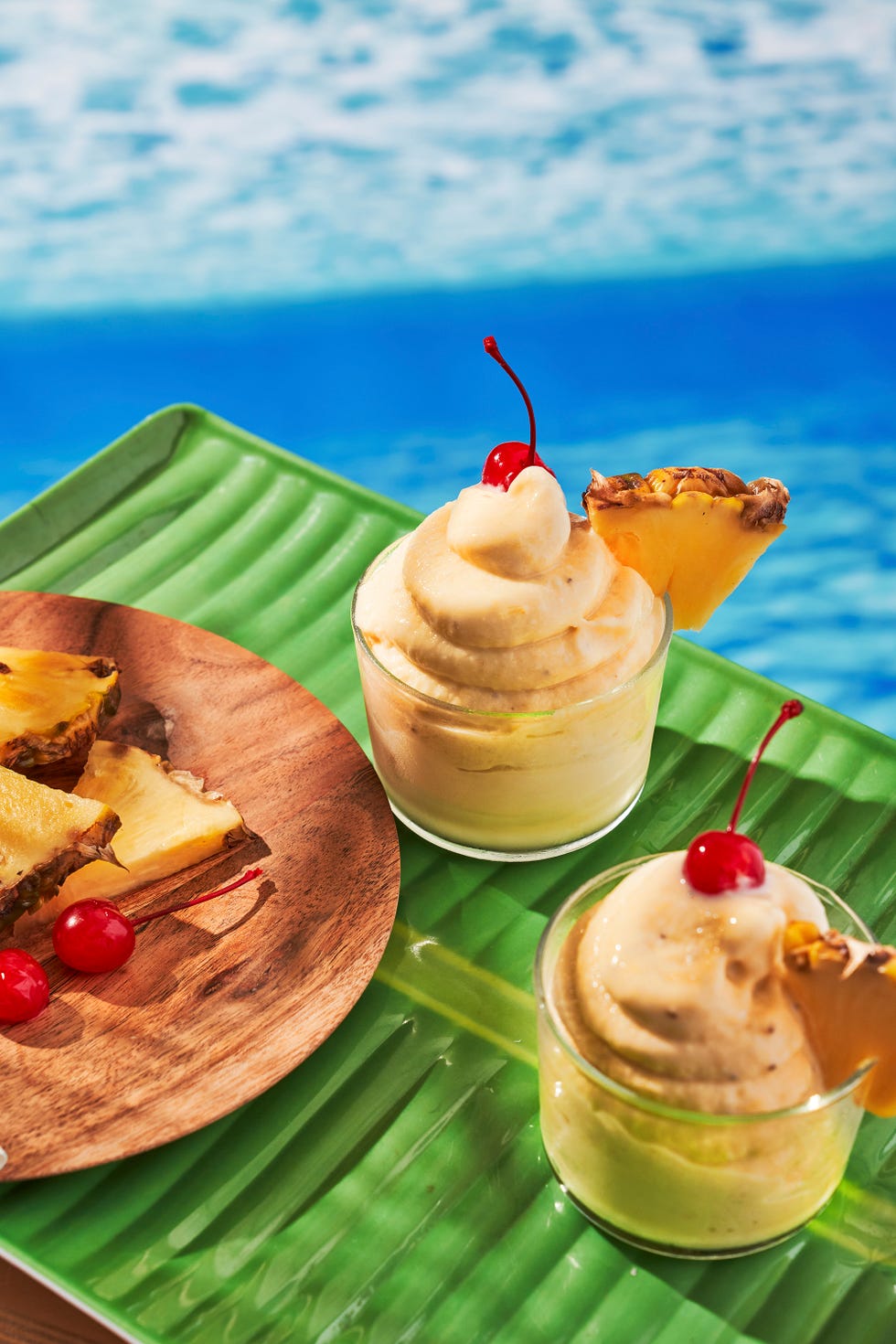 two glasses of dole whip garnished with pineapple slices and maraschino cherries on a green tray \