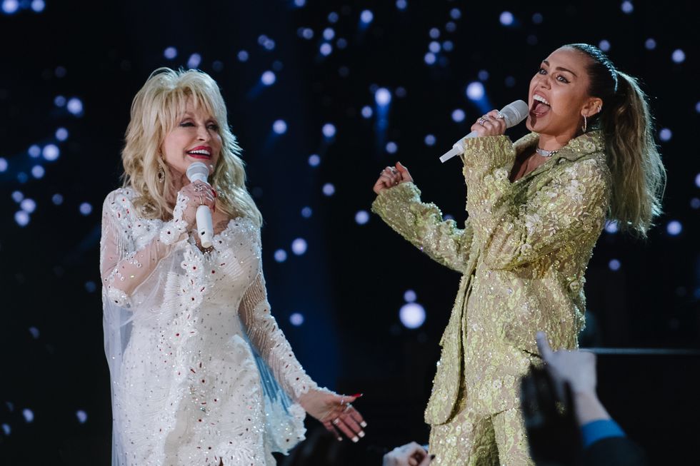 dolly parton, wearing a white dress, and miley cyrus, wearing a gold outfit, as both sing into handheld microphones on a stage, with blue lights behind them