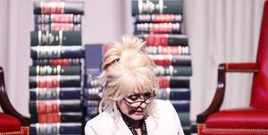 dolly parton reads her book the coat of many colors to school children, she sits on a chair in front of several stacks of books and wears a white blouse and reading glasses