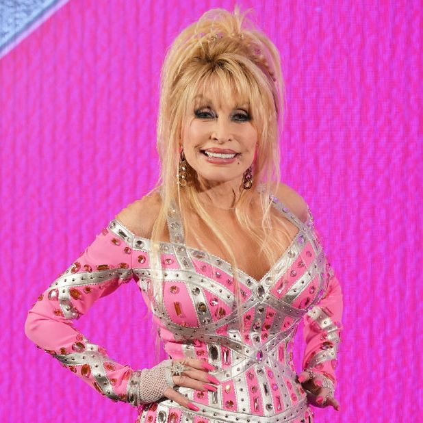 dolly parton smiles at the camera with her hands on her hips as she stands in front of a hot pink background, she wears a pink and silver bejeweled dress with gold dangling earrings