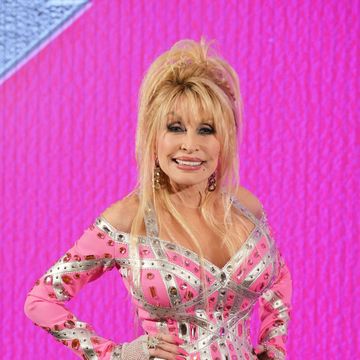 dolly parton smiles at the camera with her hands on her hips as she stands in front of a hot pink background, she wears a pink and silver bejeweled dress with gold dangling earrings