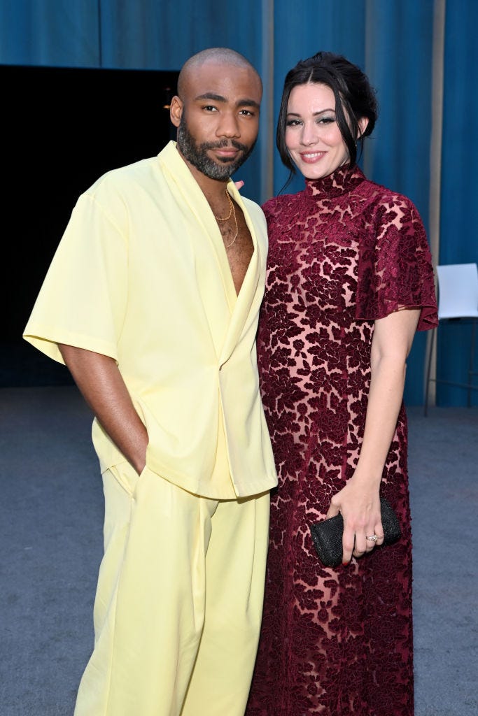 donald glover and michelle white stand together and smile for a photo, he wears a pale yellow suit, she wears a red velvet patterned short sleeve dress