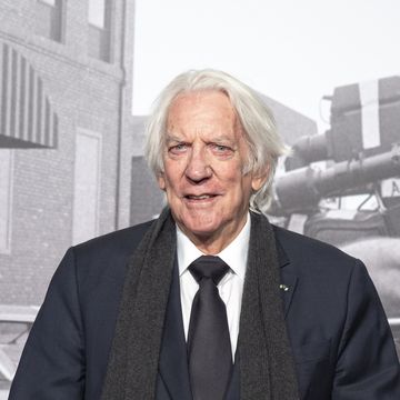 donald sutherland smiles at the camera, he wears a dark suit jacket and tie with a white dress shirt and a gray scarf