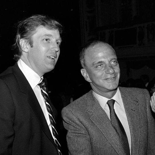 roy cohn shaking a hand as donald trump stands to his right smiling