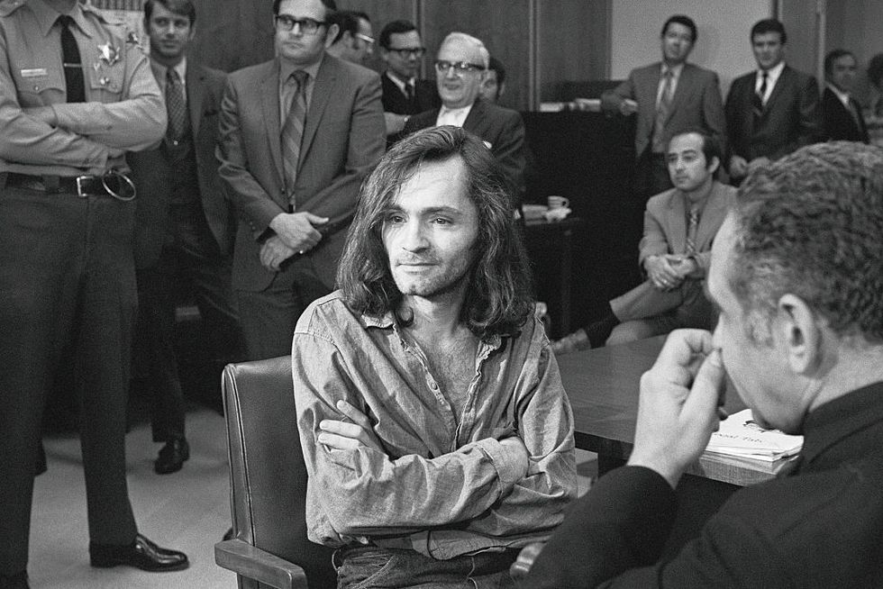 charles manson sits in court with his arms crossed, he wears a denim shirt that is partially unbuttoned and jeans, around him are several men in suits
