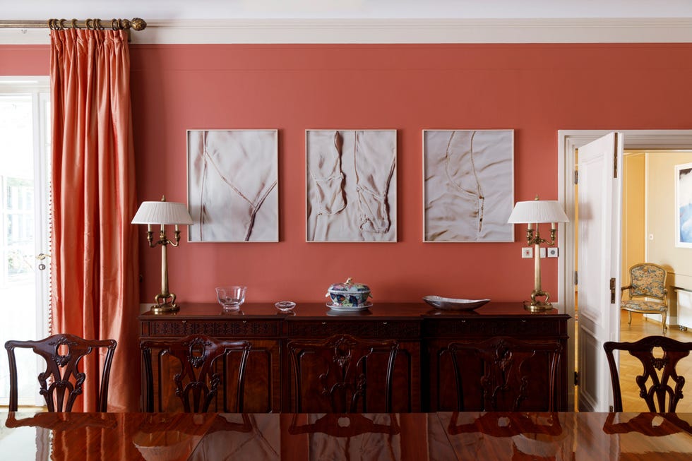 a refined dining room with a deep coral wall and three framed artworks displaying abstract patterns hang above a polished wooden sideboard