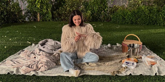 a picnic scene on grass featuring a person in a fluffy sweater surrounded by food and a picnic basket