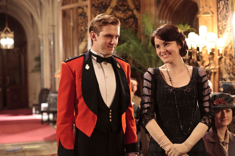 matthew crawley wearing a red military uniform walking next to lady mary wearing a black sequin evening dress inside a grand building in downton abbey