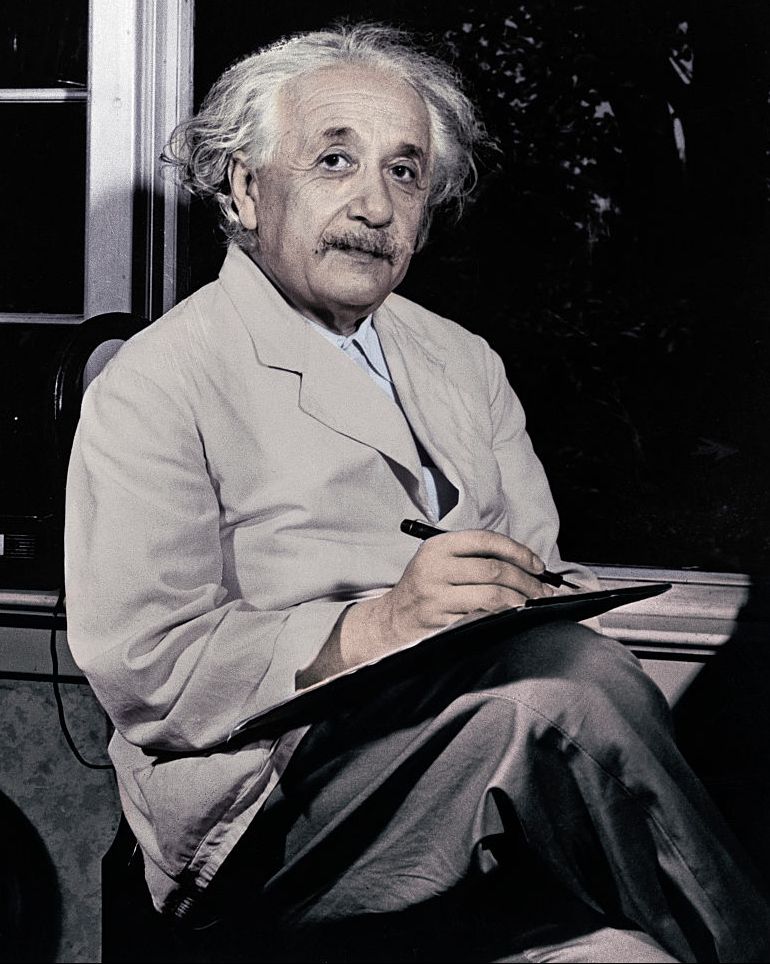 Albert Einstein sentado junto a una ventana y escribiendo en un bloc de notas mientras mira hacia arriba.
