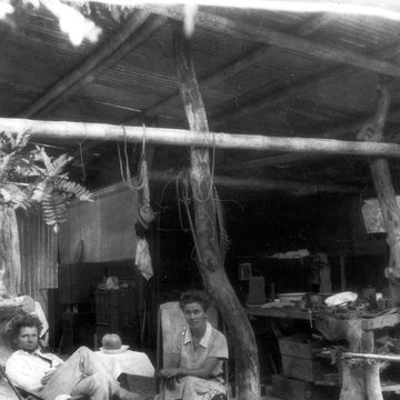 a man and a woman sit in chairs at the front of an open air shelter and look at the camera