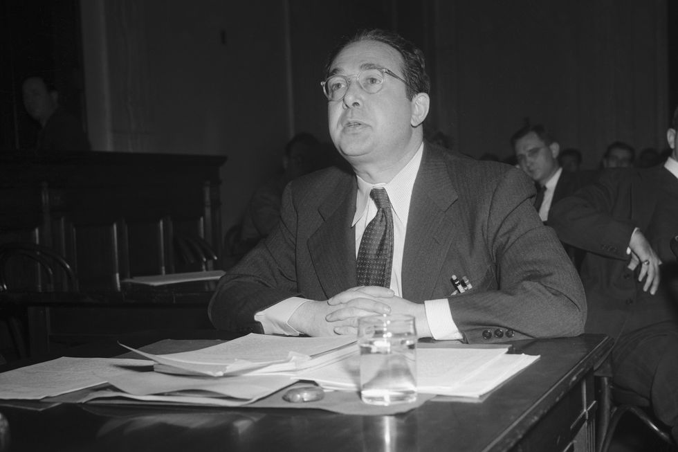 leo szilard wearing a suit and tie, sitting at a table, and speaking to someone off camera