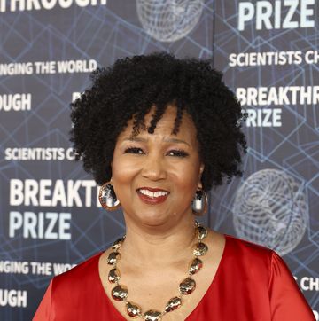 mae jemison smiles at the camera while standing in front of a photo background with designs and writing, she wears a red top with gold hoop earrings a gold necklace