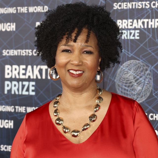 mae jemison smiles at the camera while standing in front of a photo background with designs and writing, she wears a red top with gold hoop earrings a gold necklace