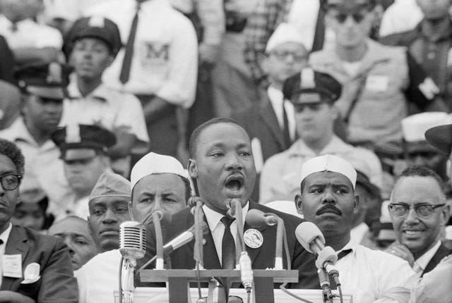 martin luther king jr speaks into several microphones in front of a lectern, he wears a suit and tie with a button on his lapel, many people watch from behind him