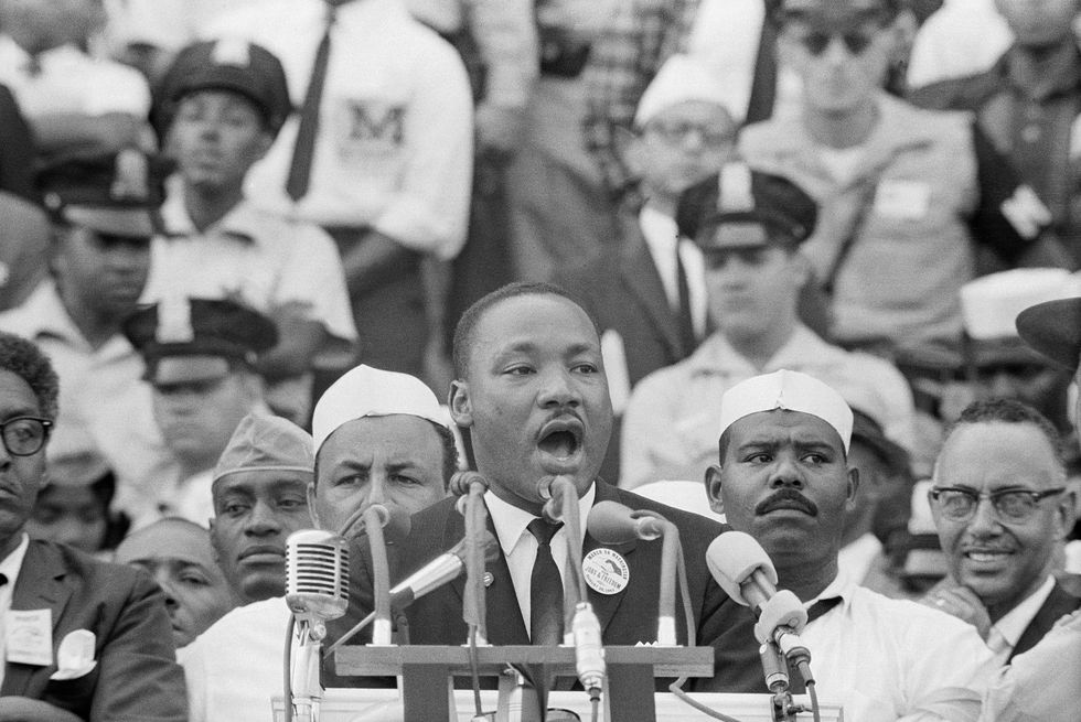 martin luther king jr speaks into several microphones in front of a lectern, he wears a suit and tie with a button on his lapel, many people watch from behind him
