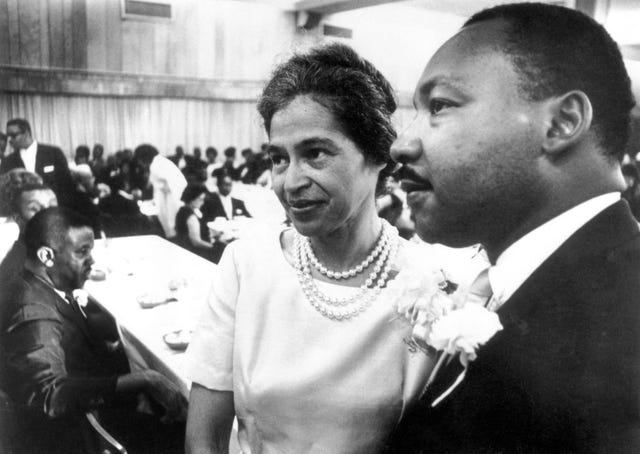 a black and white photograph of rosa parks, in a dress and pearls, and martin luther king jr, wearing a suit, standing in the foreground, with many people seated at tables in the background behind them