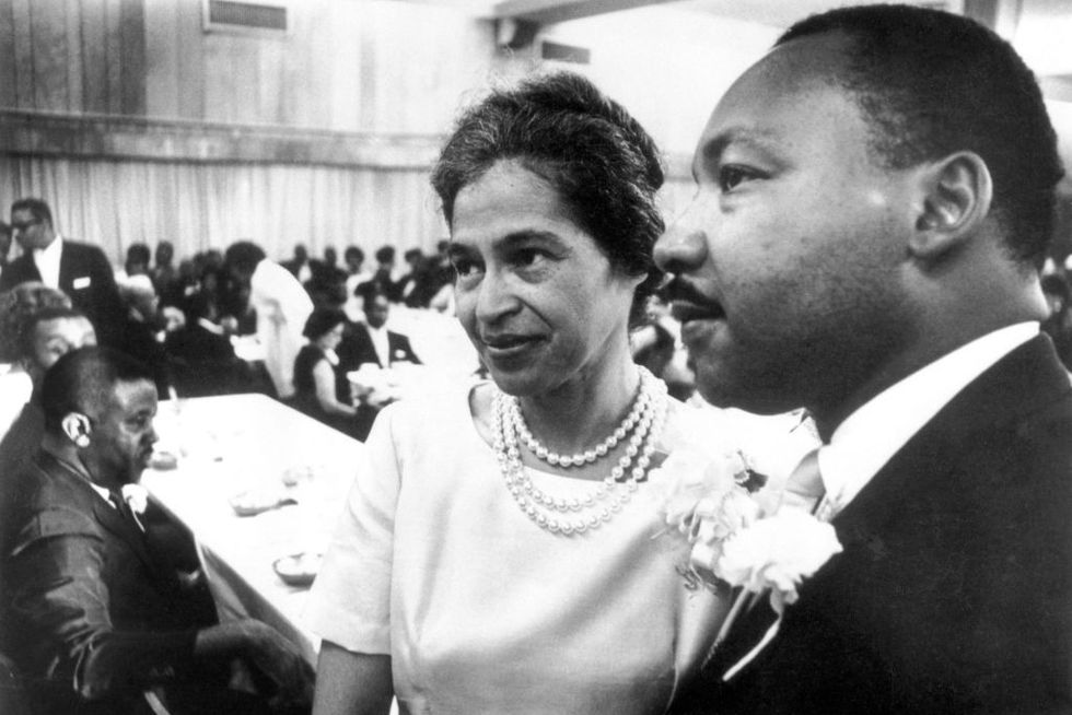 a black and white photograph of rosa parks, in a dress and pearls, and martin luther king jr, wearing a suit, standing in the foreground, with many people seated at tables in the background behind them