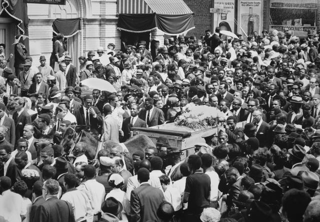 a crowd of people surround a horse drawn cart pulling a casket topped with flowers