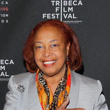 patricia bath smiles at the camera, she stands in front of a black background with white logos and wears a gray suit jacket with an orange, red, and black scarf, he holds one hand across her chest