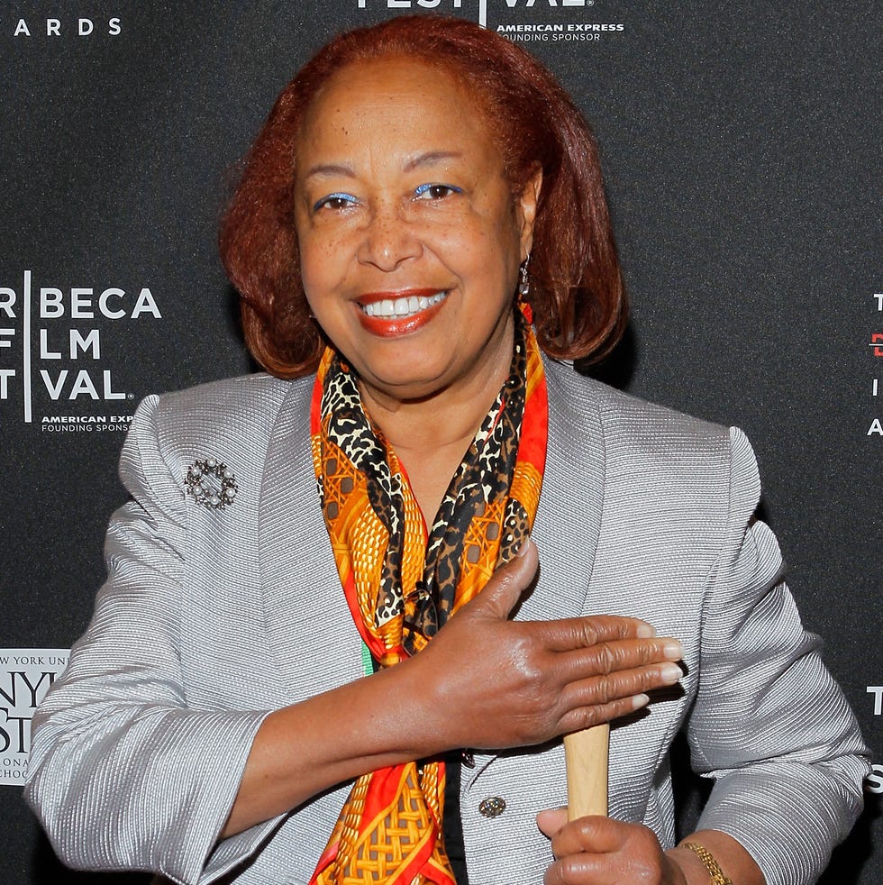 patricia bath smiles at the camera, she stands in front of a black background with white logos and wears a gray suit jacket with an orange, red, and black scarf, he holds one hand across her chest