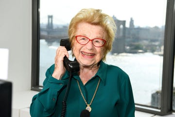 dr ruth westheimer smiles at the camera as she holds a black telephone to her ear in one hand, she wears a teal dress shirt with a gold pendant necklace