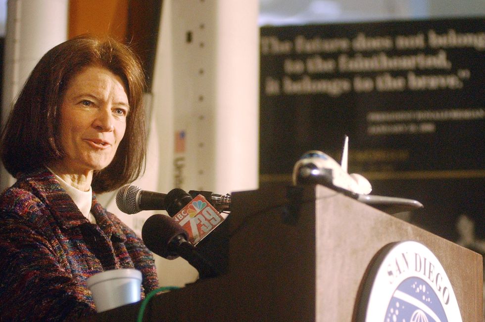 a woman in a multicolored tweed jacket speaks into microphones attached to a wooden podium