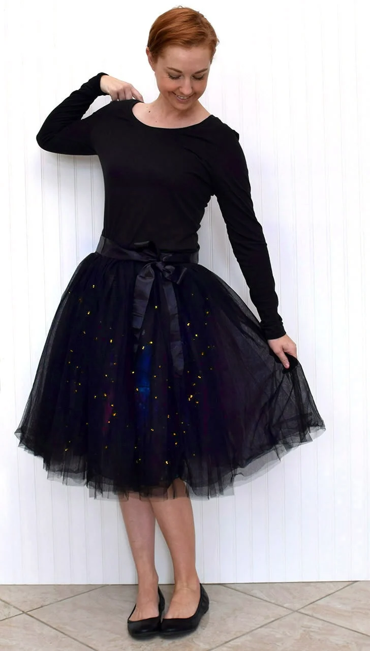 woman whith short red hair wearing a tight fitting long sleeve black shirt and a black tulle skirt with lights shining through it there is a black bow at her waist