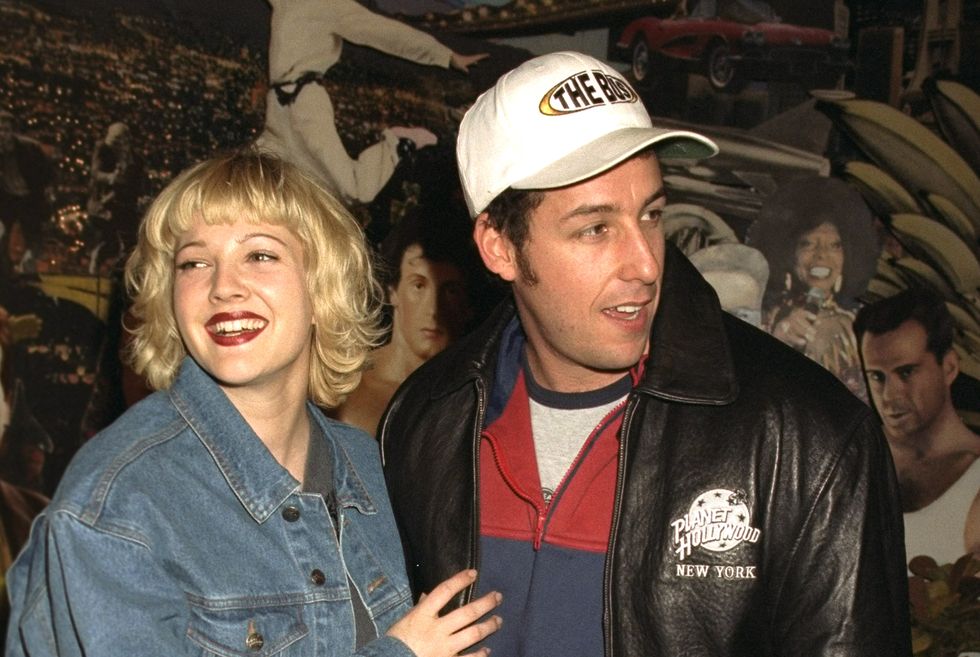 drew barrymore in a denim jacket stands next to adam sandler with her hand on his chest, she smiles and looks left of the camera, sandler looks right of the camera, he wears a black leather jacket with a planet hollywood logo a white baseball cap and a red and blue zipup jacket