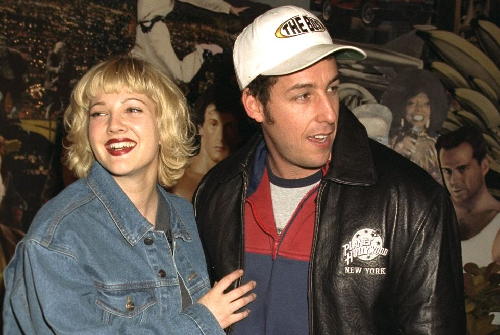 drew barrymore in a denim jacket stands next to adam sandler with her hand on his chest, she smiles and looks left of the camera, sandler looks right of the camera, he wears a black leather jacket with a planet hollywood logo a white baseball cap and a red and blue zipup jacket