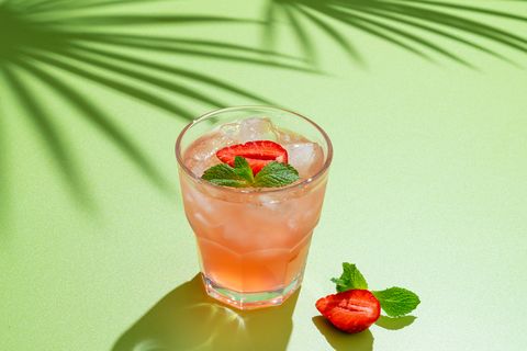drinking glass of strawberry cocktail with mint leaves and iced cubes on bright green background, tropical tree shadow homemade ready to eat lemonade vitamin delicious refreshing drink, horizontal, copy space
