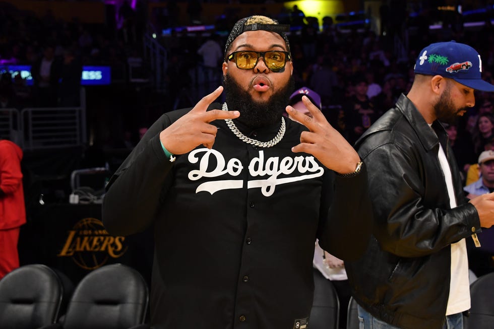 druski looks at the camera with both hands raised in peace signs, he wears a black dodgers jersey with yellow tinted glasses, a silver chain necklace and a black backwards baseball cap