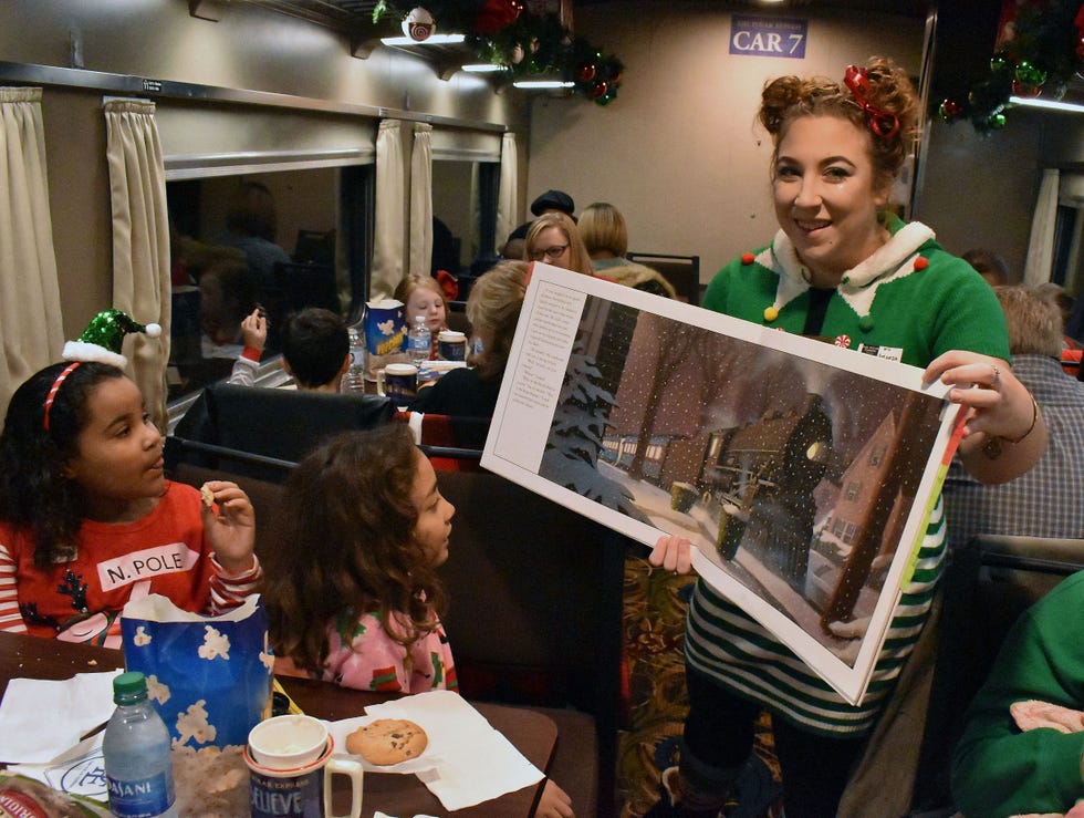 woman on a train dressed as an elf in green and white holding up an open polar express book to show two little girls sitting at a table, with more kids at tables in the background