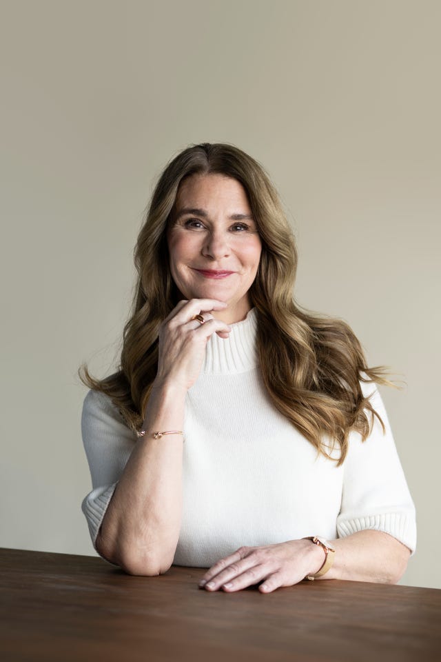 a woman with long wavy hair seated at a table wearing a white sweater