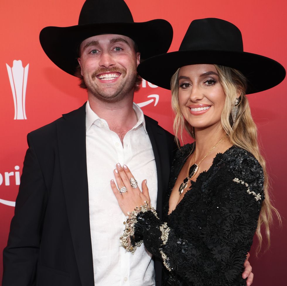 a man and woman embrace while smiling at the camera, both wear black outfits and black cowboy hats, the woman rests one hand on the man's chest