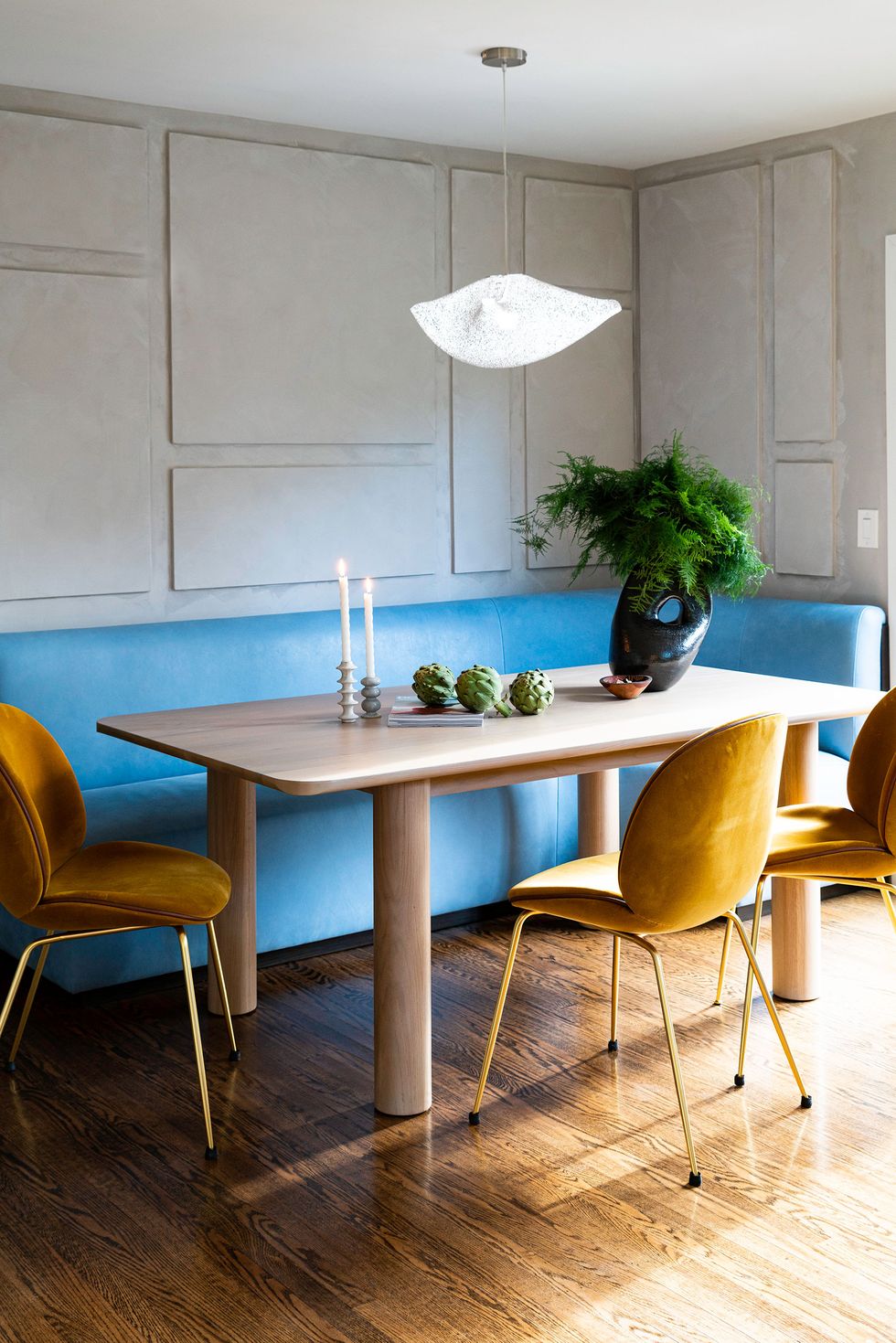 wooden dining table with rounded edges and upholstered yellow chairs with gold legs and a wraparound banquette in a bright blue