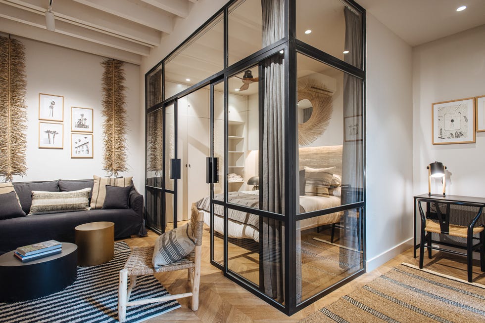 salón decorado en madera y fibras naturales junto a un dormitorio con cerramiento de vidrio con perfilería metálica