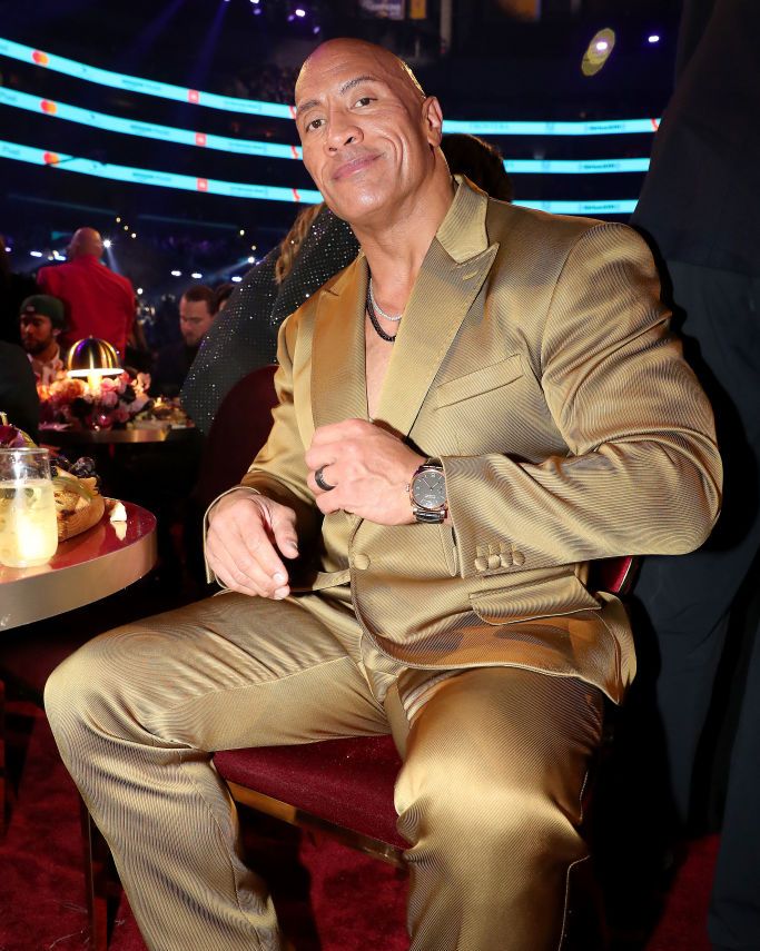dwayne johnson sitting at his table at the grammy awards and wearing a gold suit