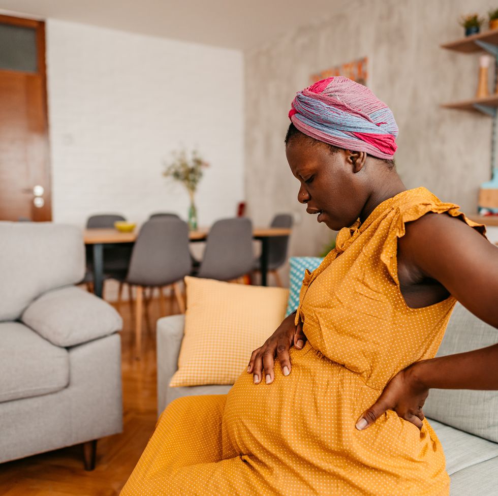 young black pregnant woman sitting on sofa and holding hands on back and stomach