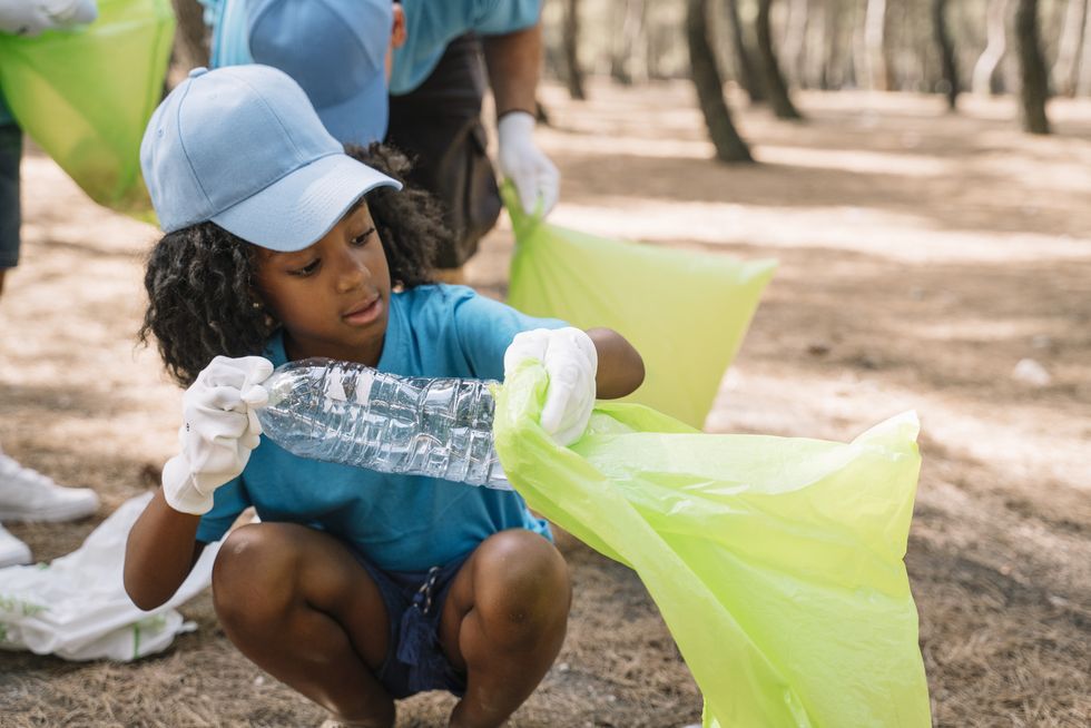 young girl in blue ballcap and tee putting plastic bottle in garbage bag in a park with other kids behind her collecting garbage