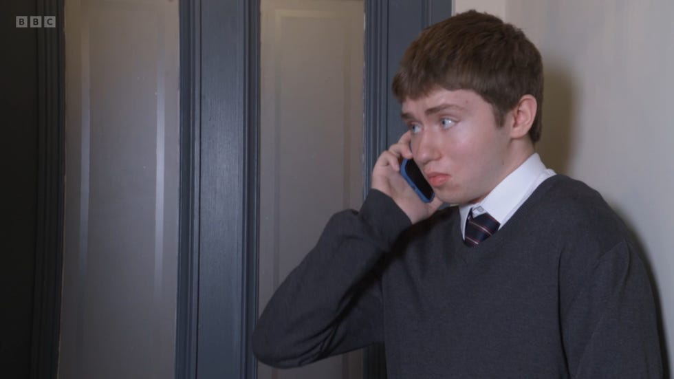 eastenders, barney in his grey school uniform, making a phone call while stood next to a door in his house