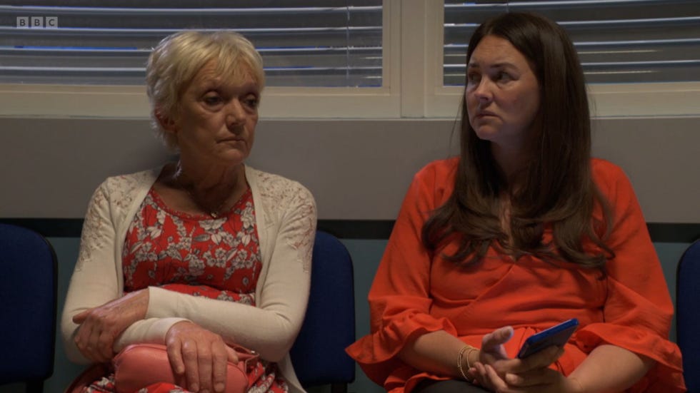eastenders, jean and stacey slater sitting in a hospital corridor, jean in a white cardigan over a floral top, and stacey in an orange blouse