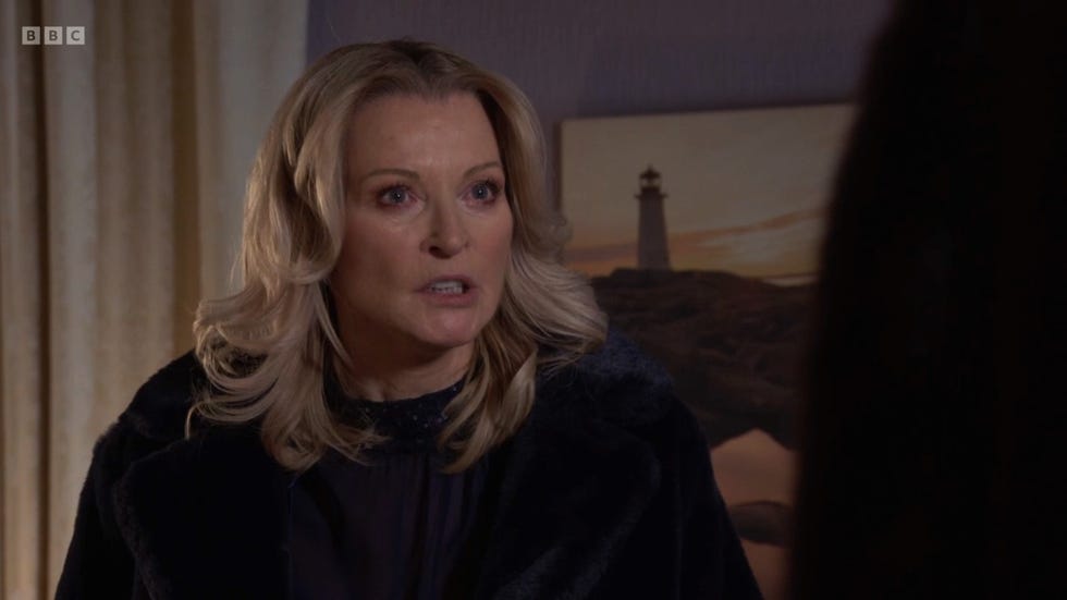 eastenders kathy beale wearing a dark coat stands with a lighthouse painting in the background