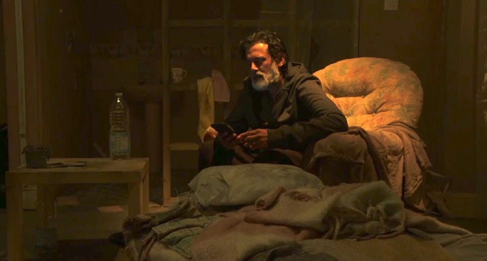 nish panesar in eastenders, a man sits in a chair while reading on a phone in a dimly lit cluttered room