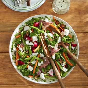 snap pea salad with feta, almonds, and prosciutto in a large white bowl with wooden serving utensils