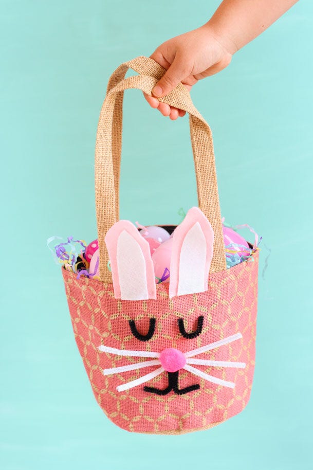 easter basket ideas, bunny basket easter basket ideas, hand holding a diy bunny basket with a pom pom nose and pipe cleaner whiskers