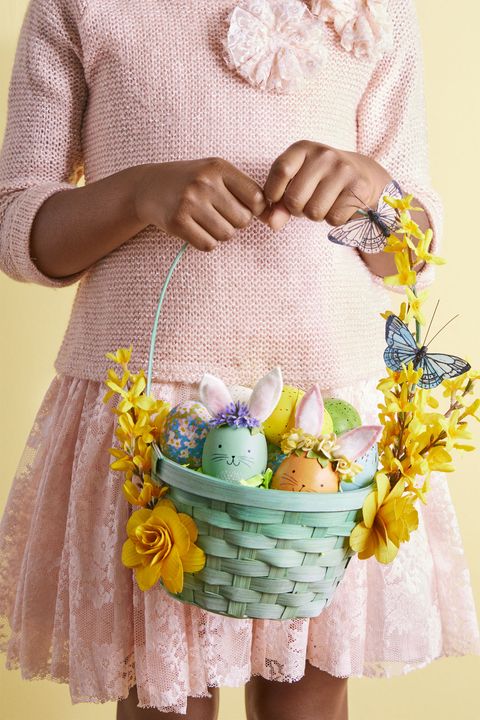 easter basket ideas, woman holding a green basket with faux flowers and butterflies on the handle, full of bunny designed eggs